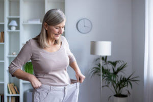 Smiling senior woman feels accomplished as she admires her weight loss progress in her large pants.